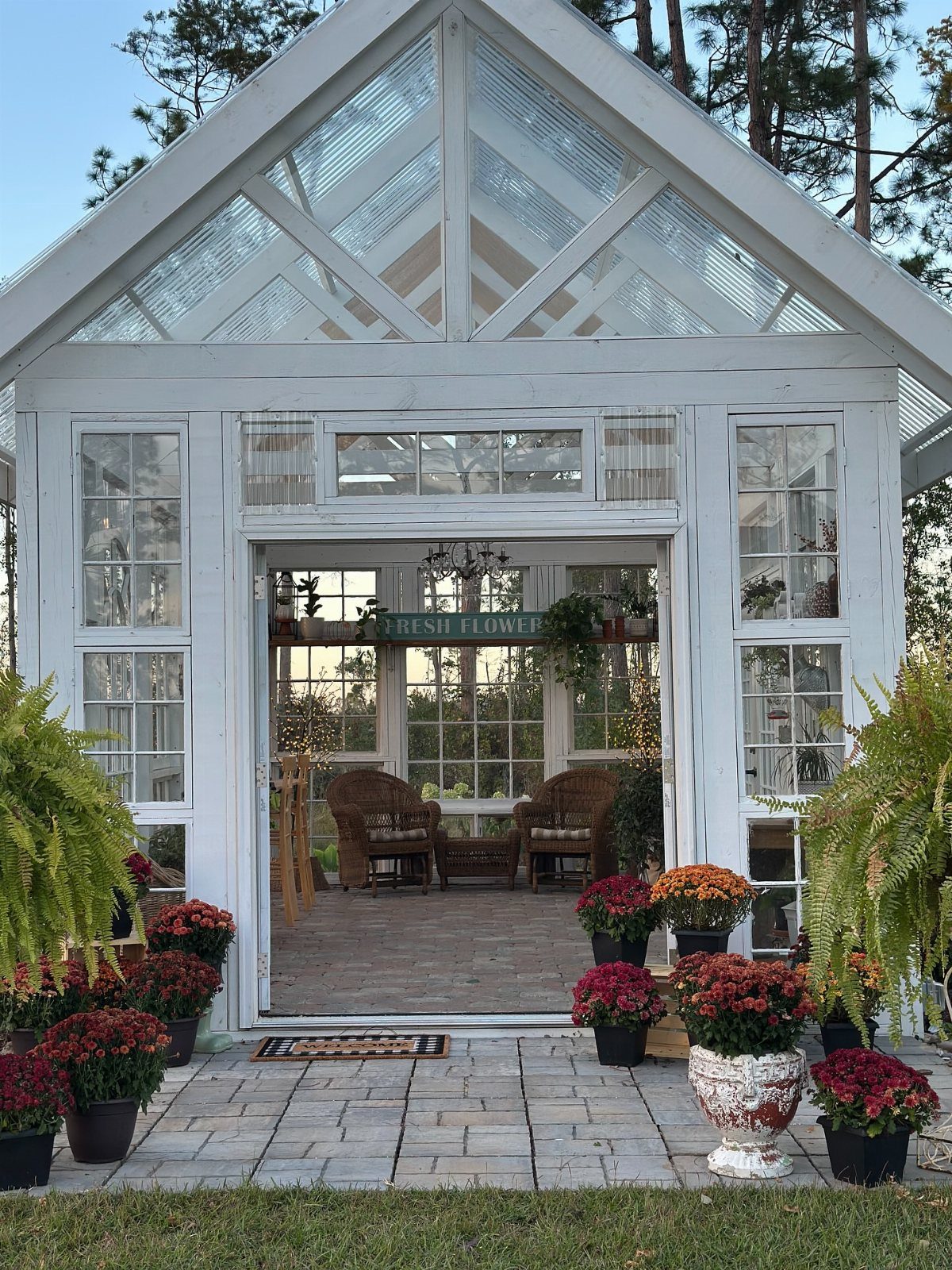White wooden greenhouse with glass roof and wicker chairs on a stone patio.